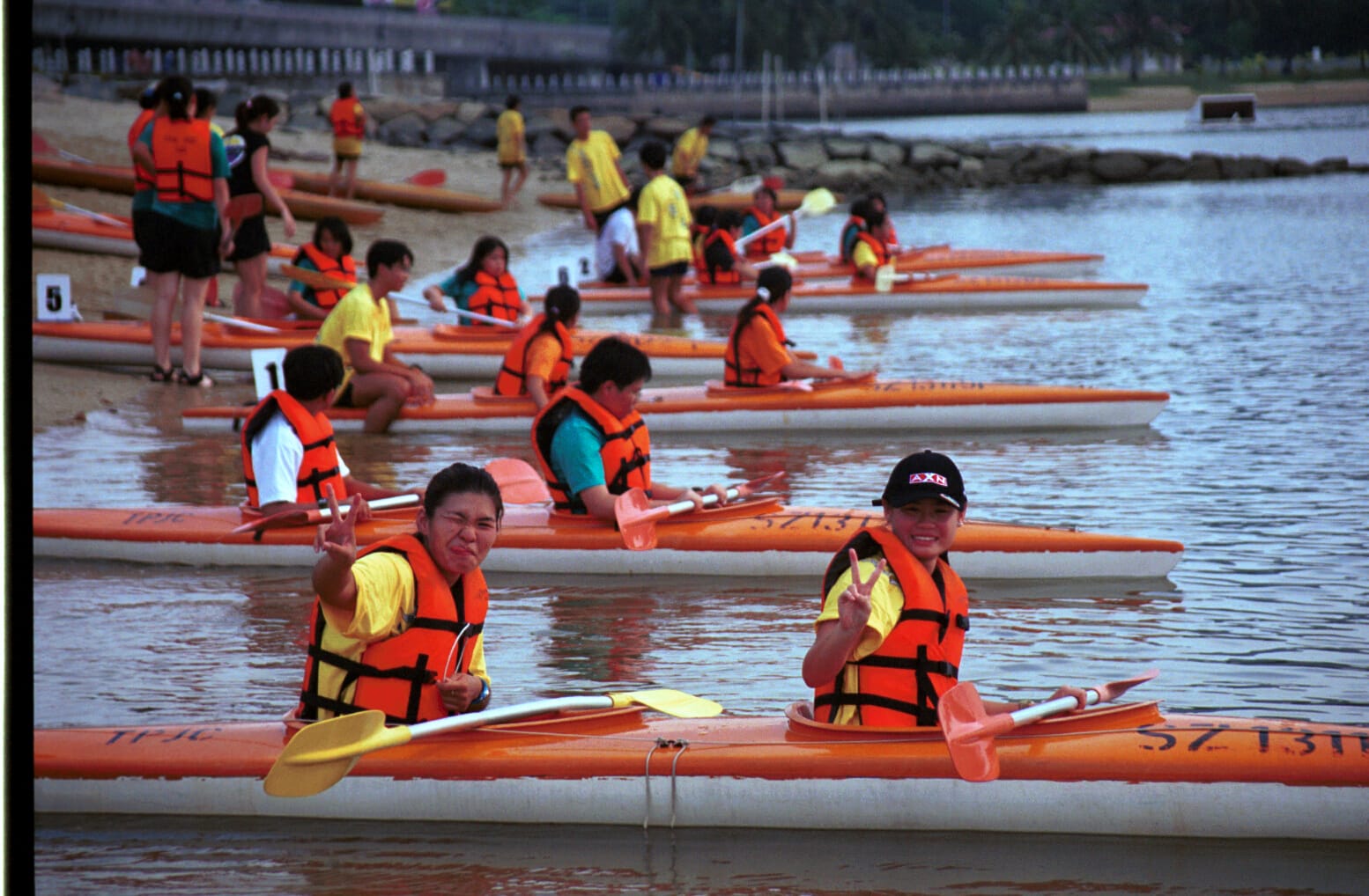 Children in orange life jackets sit in orange kayaks on calm water near a sandy beach, two posing with peace signs.